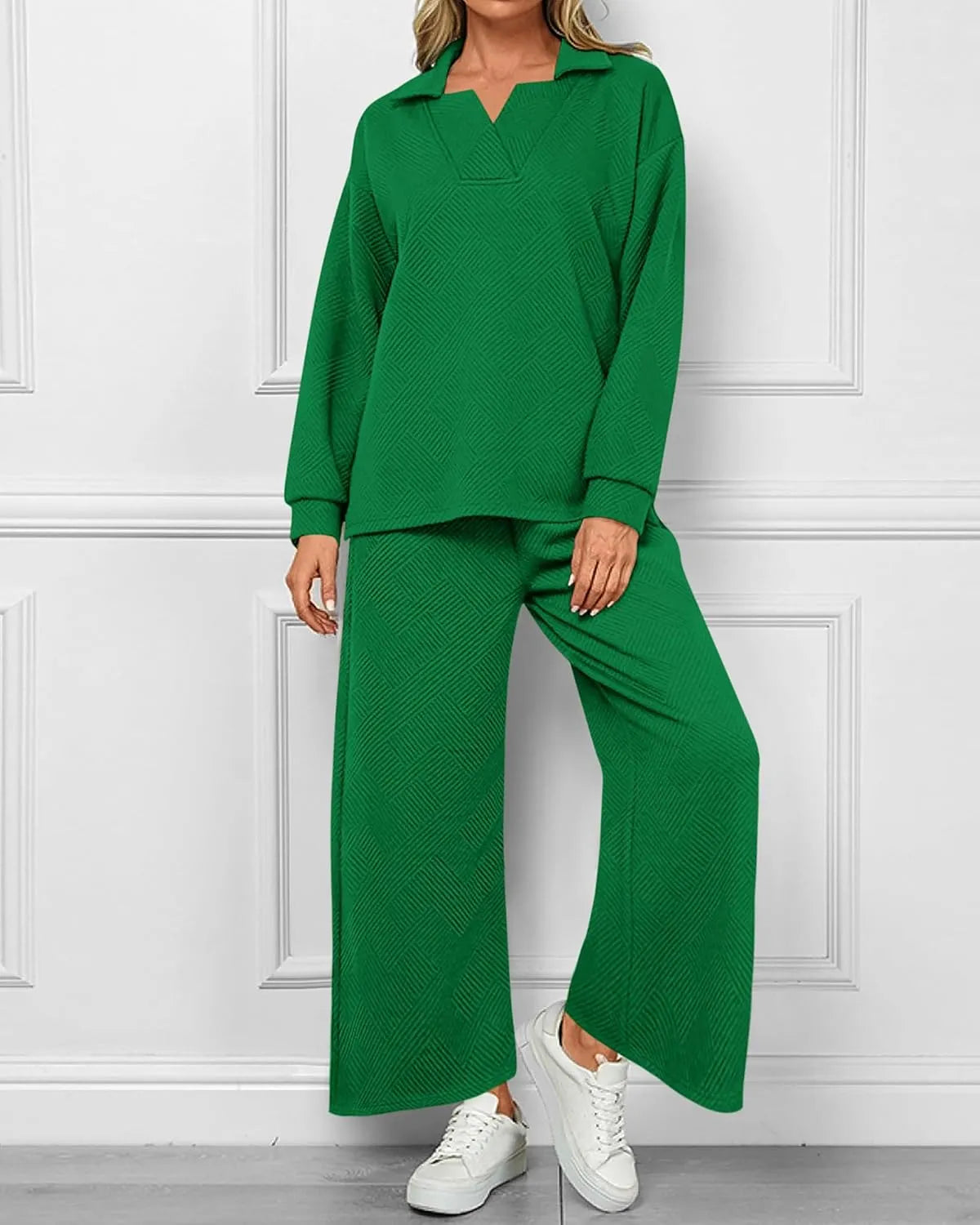 A woman modeling a matching green two-piece lounge set consisting of a long-sleeve collared top and wide-leg pants, paired with white sneakers, standing against a white paneled wall.