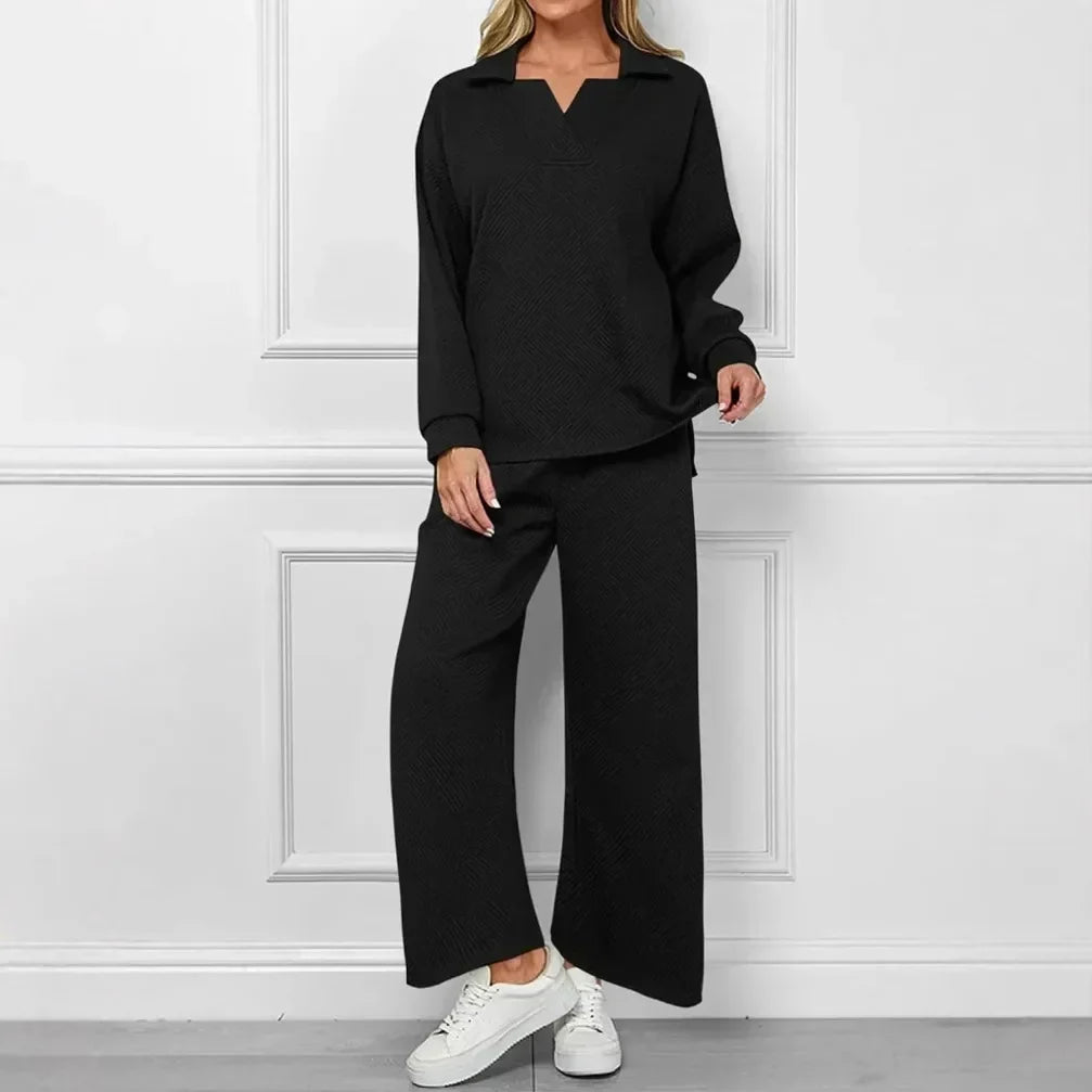 A woman modeling a black two-piece lounge set consisting of a long-sleeve collared top and matching wide-leg pants, paired with white sneakers, standing against a white paneled wall.