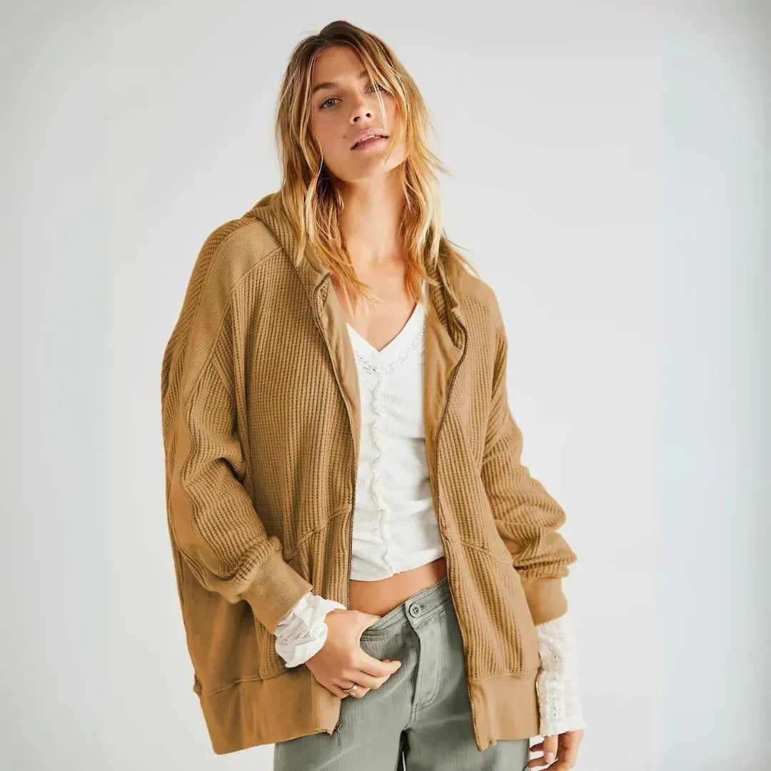 A woman wearing a tan ribbed knit zip-up hoodie with white lace-trimmed sleeves, styled over a white crop top and gray jeans, photographed against a light gray studio background.