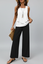 Woman wearing a white sleeveless top and black wide-leg pants against a white wall.