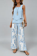 A woman models a two-piece matching set featuring a light blue sleeveless top with a ruffled layered hem and wide-leg blue and cream leaf-patterned pants, styled with accessories in an outdoor patio setting.