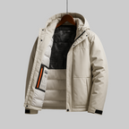 A light beige hooded puffer jacket hanging on a wooden hanger against a solid gray background. The jacket is shown open, revealing a black mesh-lined interior and a zippered pocket on the left side with a small orange and black label.