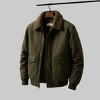 An olive green bomber jacket with a brown sherpa collar, hanging on a wooden hanger against a solid gray background.