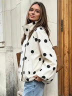 A woman wearing a cream-colored puffer jacket with large black polka dots, paired with light blue jeans and a brown shoulder bag, standing outdoors next to a light-colored stone wall and a wooden door.