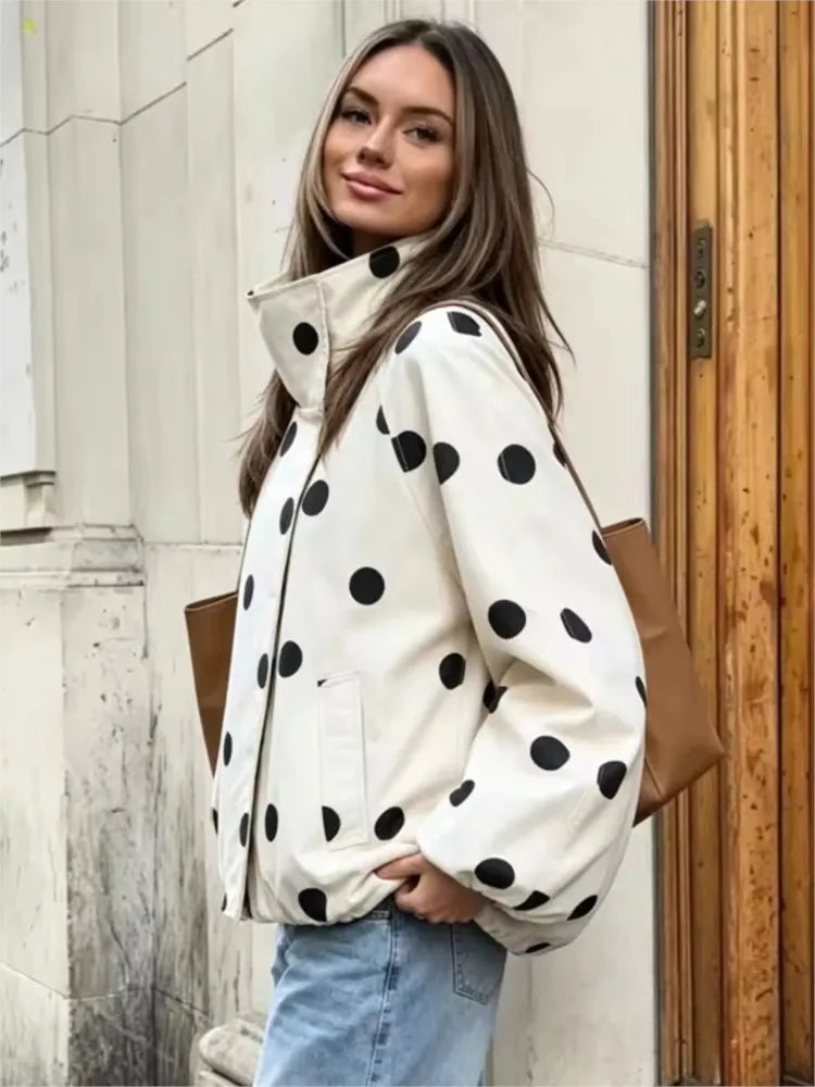 A woman wearing a cream-colored puffer jacket with large black polka dots, paired with light blue jeans and a brown shoulder bag, standing outdoors next to a light-colored stone wall and a wooden door.