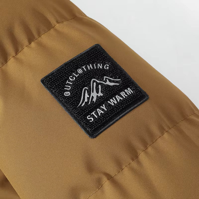 Close-up of a black rectangular embroidered patch on a tan puffer jacket. The patch features white text reading 'OUTCLOTHING' and 'STAY WARM' with a white mountain graphic in the center.