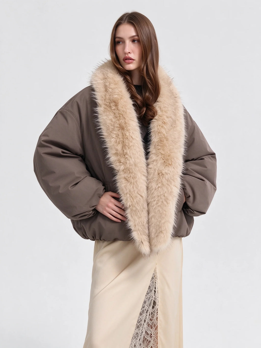 A model wearing a voluminous taupe-brown puffer jacket with a large beige faux-fur collar, paired with a cream skirt, against a plain light gray studio background.