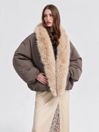 A model wearing a voluminous taupe-brown puffer jacket with a large beige faux-fur collar, paired with a cream skirt, against a plain light gray studio background.