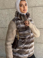 Woman wearing a fur vest over a beige sweater against a stone wall.