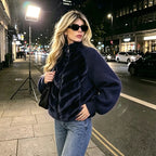 A woman posing on a city street at night wearing a navy blue textured zip-up jacket with ruffle details, blue jeans, and black sunglasses.