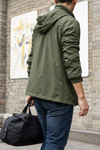 A man wearing an olive green hooded windbreaker jacket, shown from the back as he walks against a brick wall background while carrying a black duffel bag.