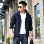 A man wearing a dark navy lightweight zip-up jacket over a white t-shirt and jeans, standing outdoors against a brick wall background.