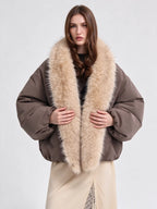 A model wearing a brown puffer jacket with a large beige fur collar, styled with a cream skirt against a light gray studio background.