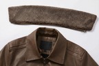 Close-up detail shot of a brown leather jacket with a matching brown sherpa fleece neck warmer, showing the collar, zipper, and interior brand label 'Katie Seth'.
