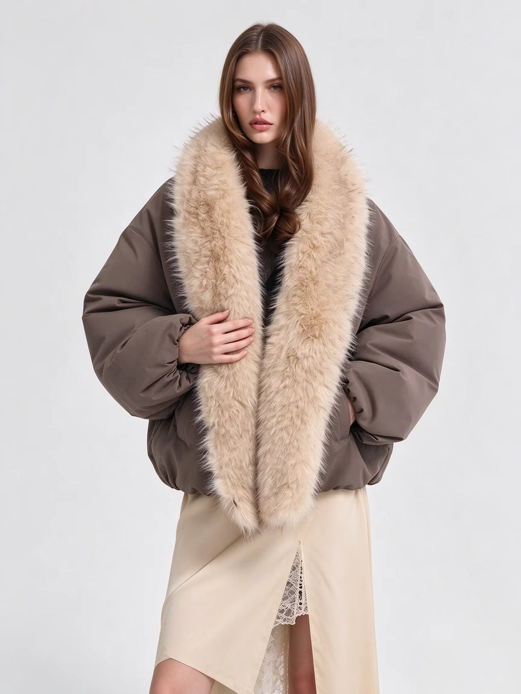 A model wearing a brown puffer jacket with a large beige faux fur collar, styled with a beige wrap skirt against a light gray studio background.