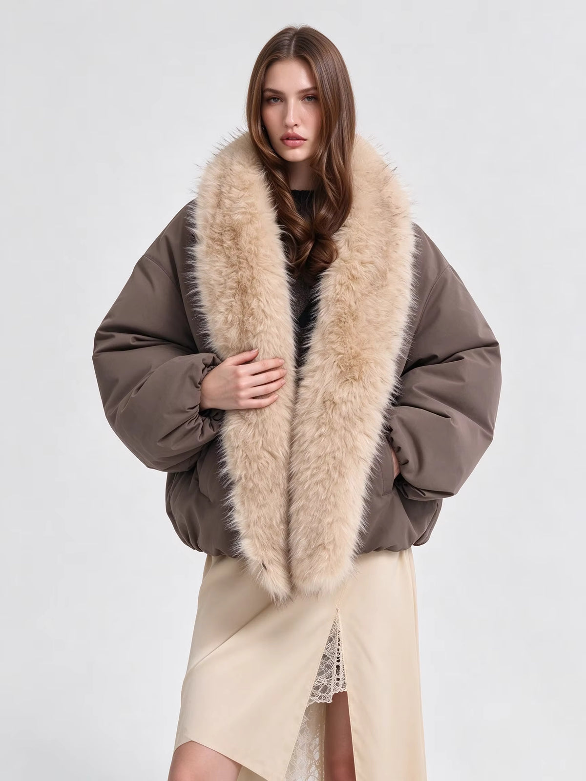 A model wearing a brown puffer jacket with a large beige faux fur collar, styled with a beige wrap skirt against a light gray studio background.
