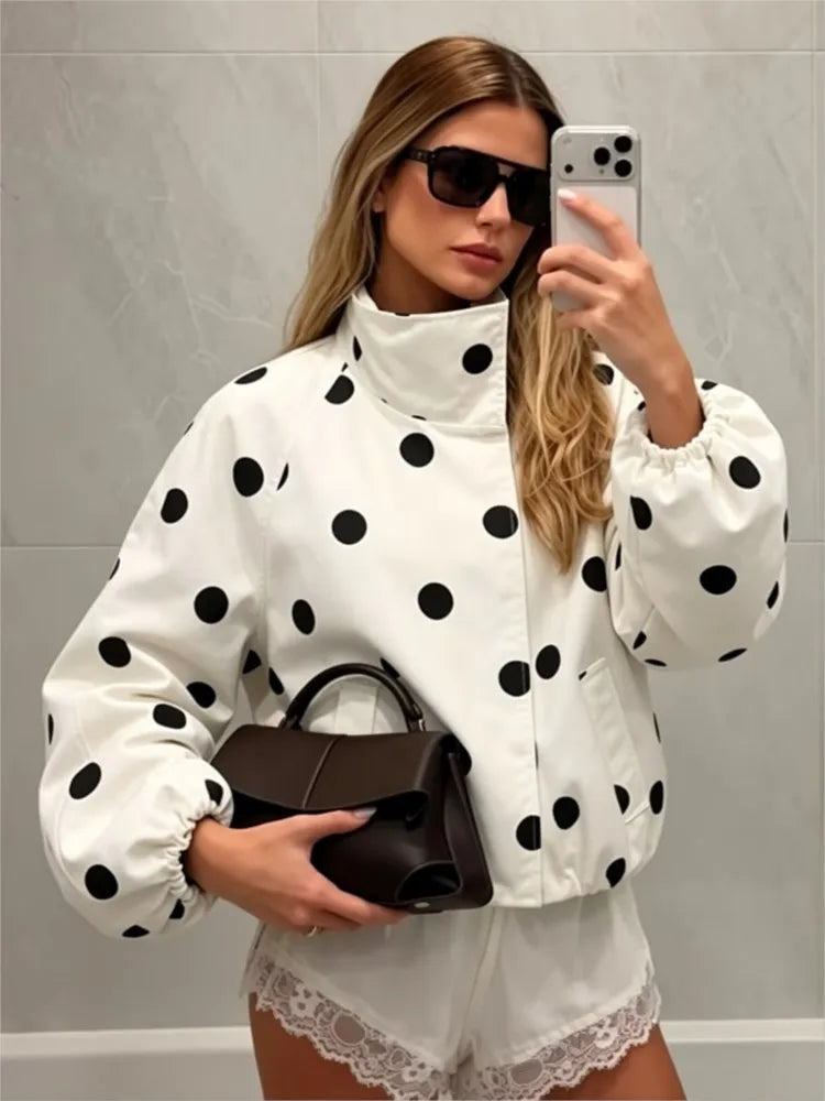 A woman takes a mirror selfie wearing a white puffer jacket with black polka dots, white lace-trimmed shorts, black sunglasses, and holding a dark brown handbag.