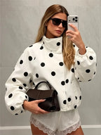 A woman takes a mirror selfie wearing a white puffer jacket with black polka dots, white lace-trimmed shorts, black sunglasses, and holding a dark brown handbag.