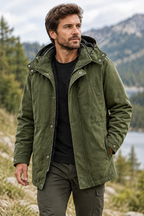 A man wearing an olive green hooded jacket over a black sweater and dark pants, standing outdoors in a mountain and lake setting.