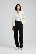 A model stands against a light gray studio background wearing a cream zip-front jacket over a white top, black wide-leg trousers, and white sneakers.