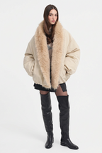 A model wearing a beige puffer jacket with a large beige fur collar, paired with a black mini skirt and black over-the-knee boots, against a plain white studio background.