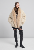 A model stands against a light gray studio background wearing a beige puffer jacket with a large beige faux-fur collar, black shorts, sheer black tights, and black over-the-knee boots.
