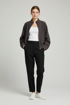 A model stands against a light gray studio background wearing a dark gray zip-front jacket, a white tank top, black trousers, and white sneakers.