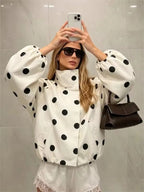 A woman takes a mirror selfie wearing a white puffer jacket with large black polka dots, white shorts, sunglasses, and carrying a dark brown shoulder bag against a light gray tiled wall.