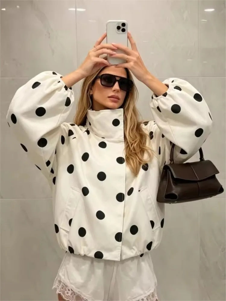 A woman takes a mirror selfie wearing a white puffer jacket with large black polka dots, white shorts, sunglasses, and carrying a dark brown shoulder bag against a light gray tiled wall.