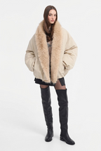A model wearing a beige puffer jacket with a large, fluffy beige faux-fur collar, styled with a black mini skirt and black knee-high boots against a plain white background.
