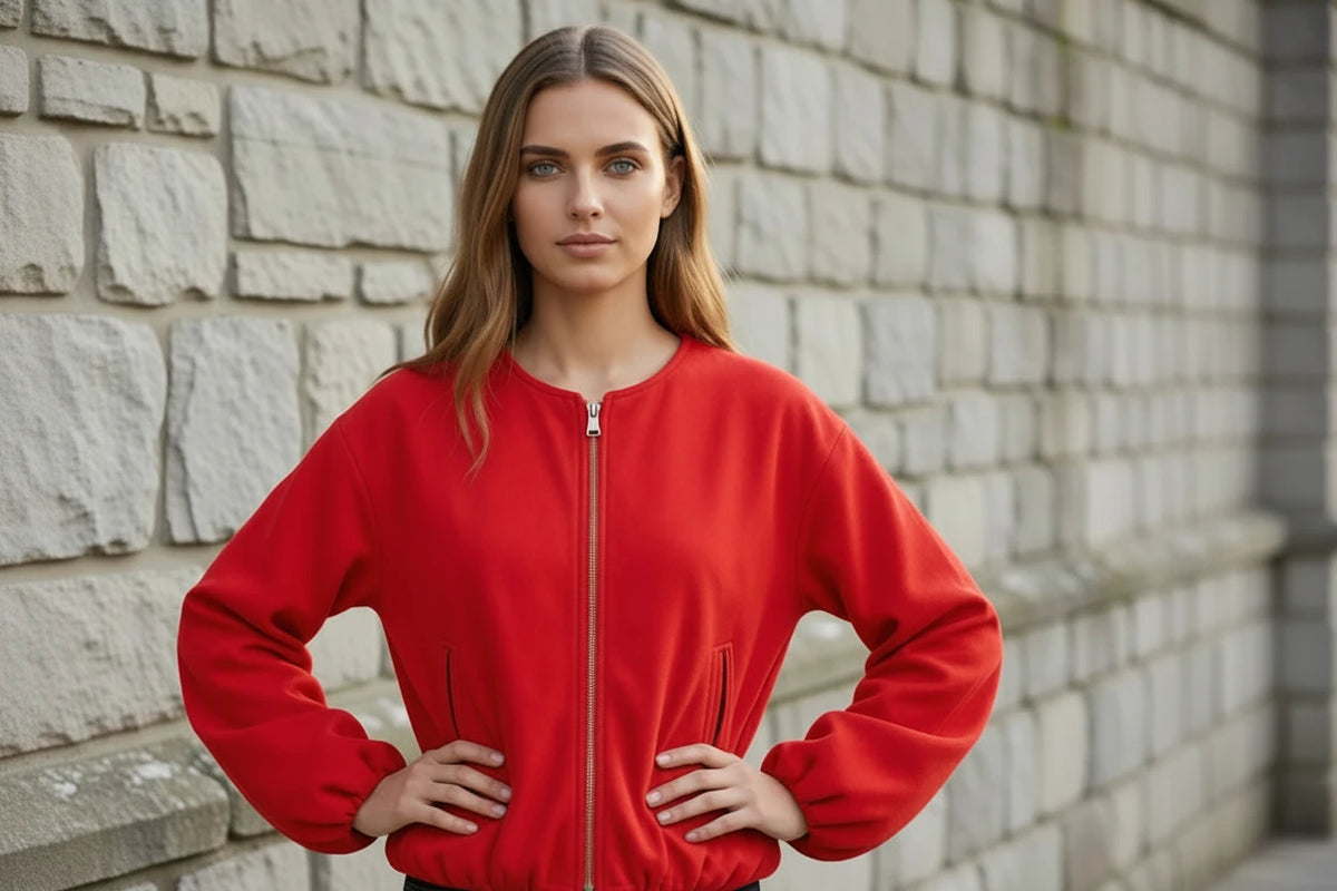 A woman wearing a bright red zip-up bomber jacket with side zip pockets, paired with dark blue jeans, standing against a light gray stone wall.
