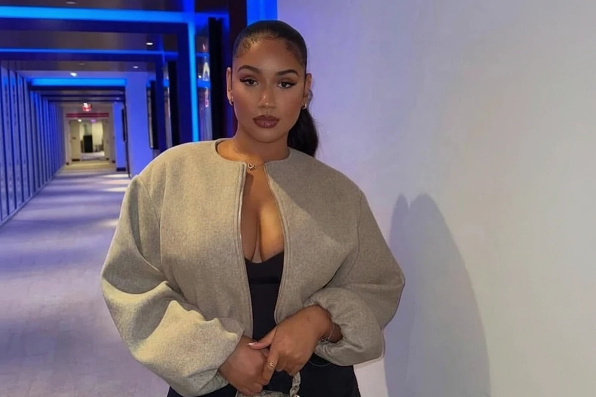 A woman wearing a light beige oversized zip-up bomber jacket over a black top and black pants, holding a beige and black patterned handbag. The background shows a hallway with blue lighting on the left and a plain wall on the right.