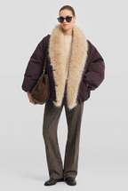 A model wearing a dark brown puffer jacket with a massive, fluffy cream-colored faux fur collar, styled with a cream ribbed sweater, brown trousers, and a brown suede shoulder bag against a light gray background.
