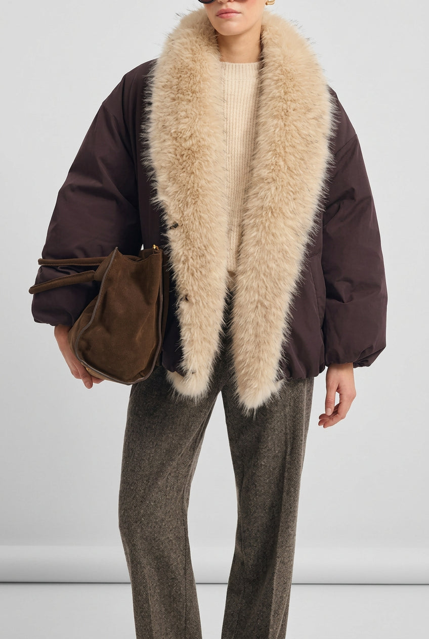 A model wearing a dark brown puffer jacket with a massive, fluffy cream-colored faux fur collar, styled with a cream ribbed sweater, brown trousers, and a brown suede shoulder bag against a light gray background.