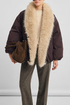 A model wearing a dark brown puffer jacket with a massive, fluffy cream-colored faux fur collar, styled with a cream ribbed sweater, brown trousers, and a brown suede shoulder bag against a light gray background.