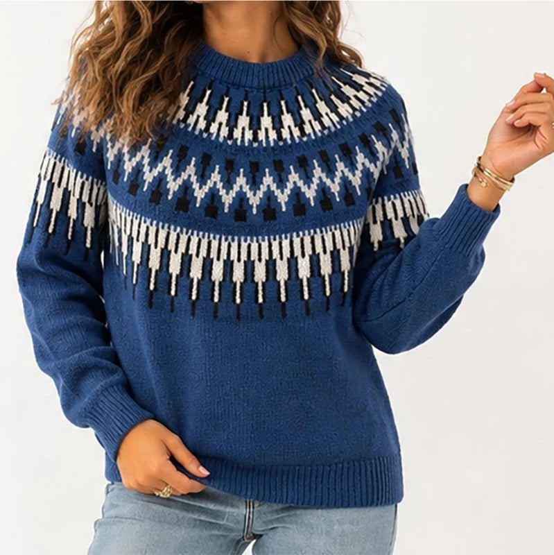 A woman wearing a blue knit sweater with a white and black geometric Fair Isle pattern across the chest and shoulders, paired with light blue jeans against a plain white background.