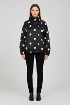 A model stands against a light gray studio background wearing a black zip-front jacket with large white polka dots, black skinny pants, and black ankle boots.