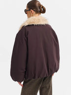 A person shown from the back wearing a dark plum-brown puffer jacket with a fluffy cream faux-fur collar, paired with brown textured trousers, against a plain light gray studio background.