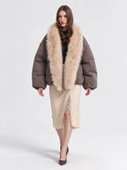 A model wearing a brown puffer jacket with a large beige fur collar, paired with a beige midi skirt and black heels, against a plain light gray studio background.