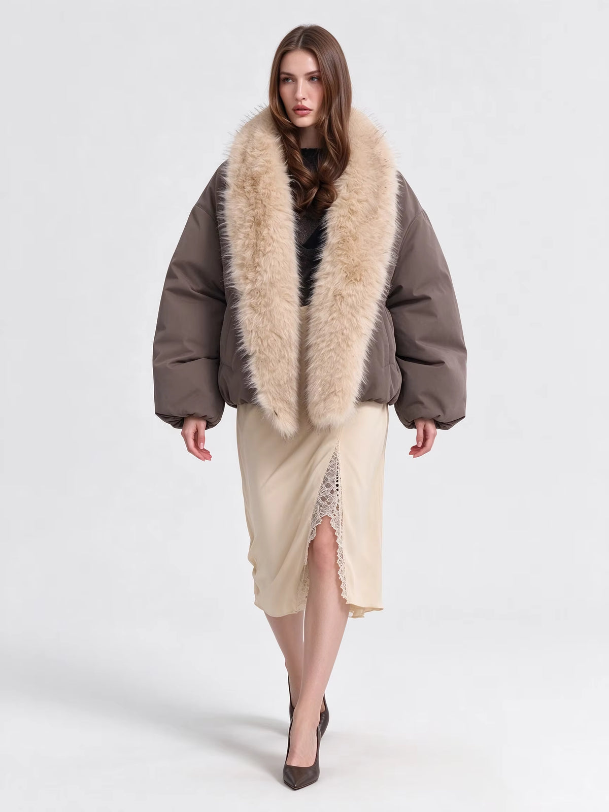 A model wearing a brown puffer jacket with a large beige fur collar, paired with a beige midi skirt and black heels, against a plain light gray studio background.