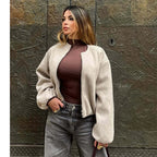 A woman wearing a beige fuzzy zip-up bomber jacket over a dark brown mock-neck top and gray jeans, standing in front of a textured bronze metal wall.