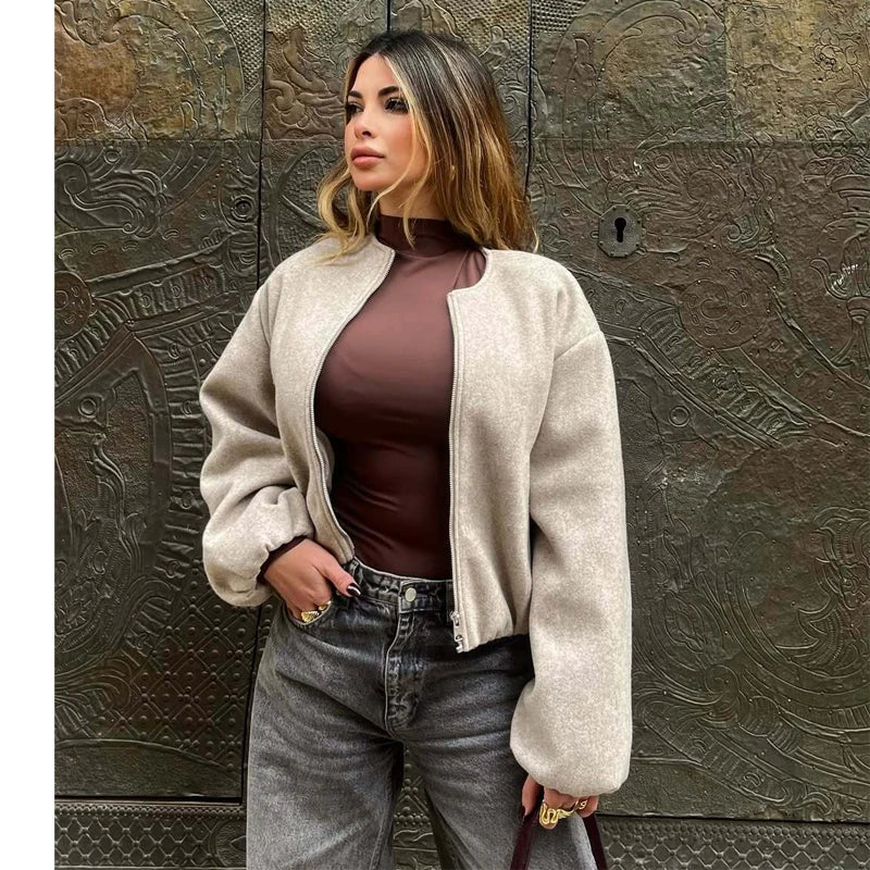 A woman wearing a beige fuzzy zip-up bomber jacket over a dark brown mock-neck top and gray jeans, standing in front of a textured bronze metal wall.