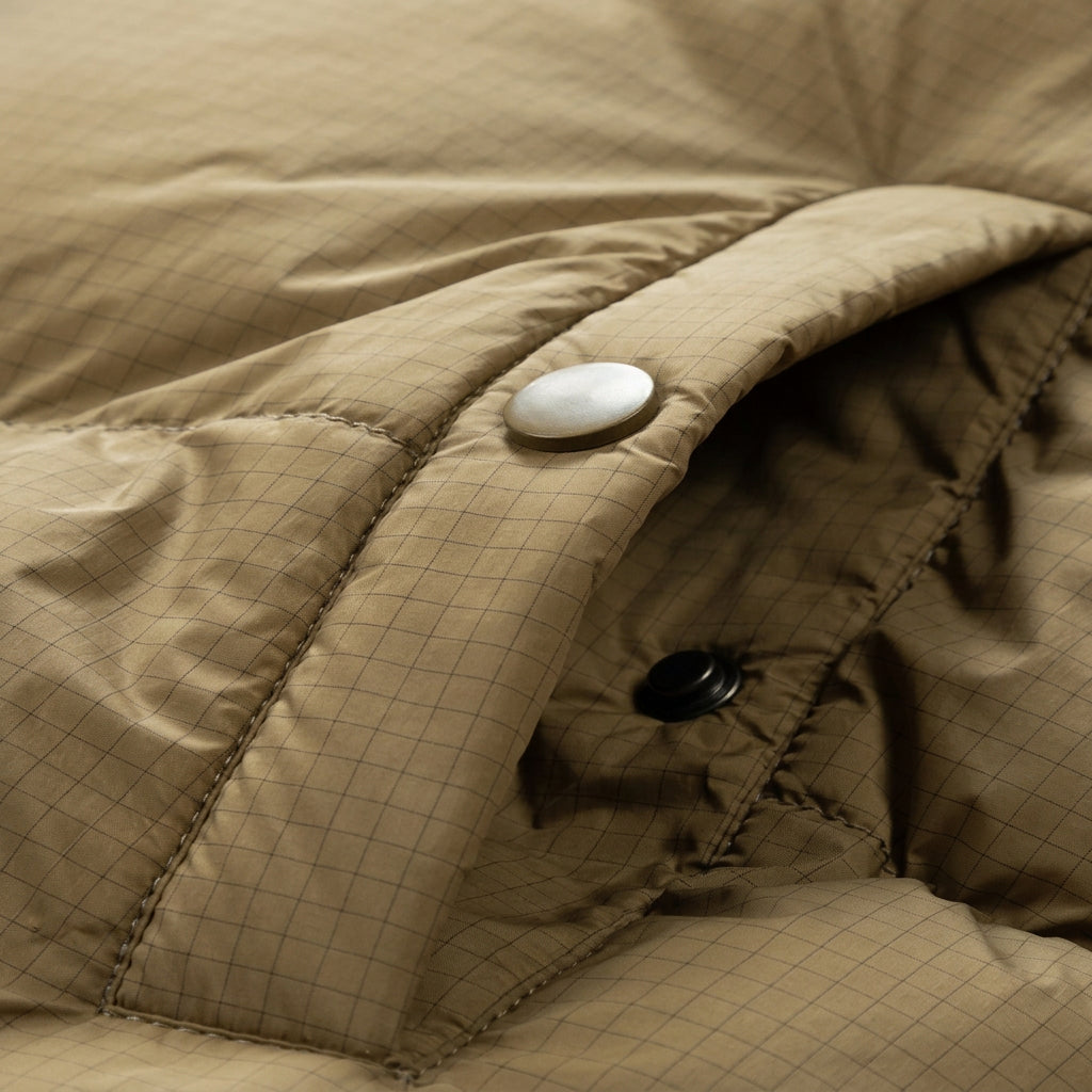 Close-up of a quilted tan jacket or puffer fabric with a side pocket, showing a white snap button, black snap button, stitched seam, and Chinese and English text at the bottom.