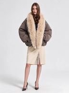 A model wearing a brown puffer jacket with a large beige fur collar, paired with a beige skirt and brown heels, photographed against a light gray studio background.
