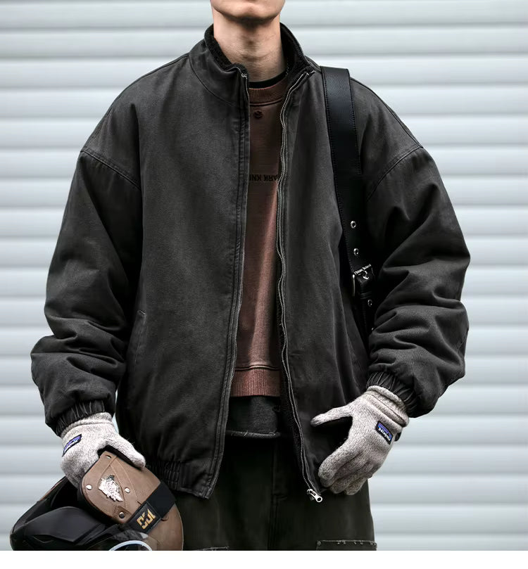 A person wearing a dark charcoal zip-front jacket over a brown shirt, with gray gloves and a black shoulder bag, standing in front of a light gray corrugated metal wall.