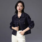 A woman modeling a dark indigo denim jacket with buttoned cuffs and front pockets, paired with white pants, against a solid gray studio background.