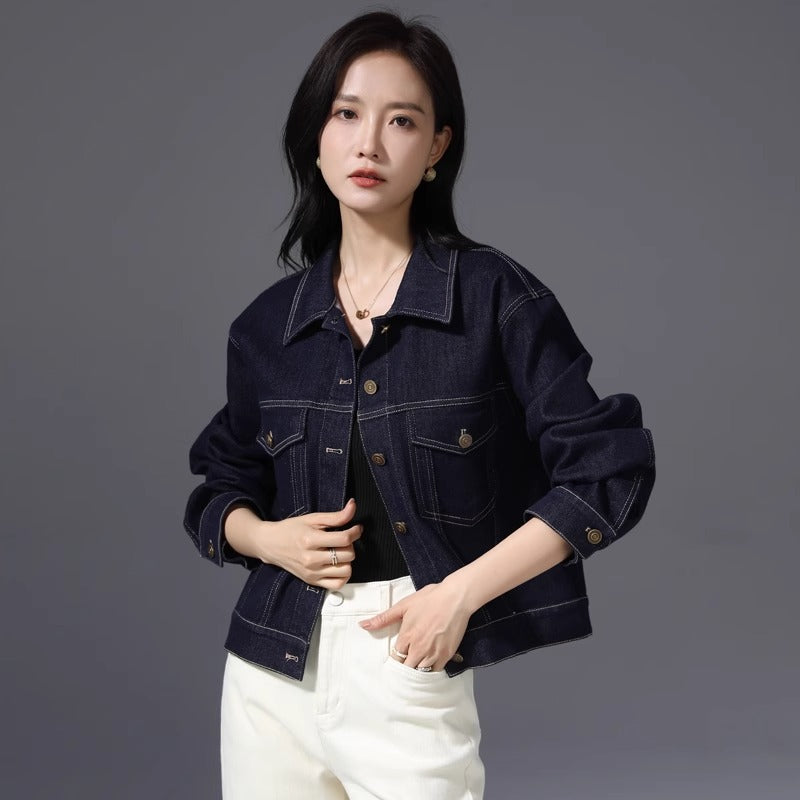 A woman modeling a dark indigo denim jacket with buttoned cuffs and front pockets, paired with white pants, against a solid gray studio background.