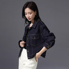 A woman wearing a dark indigo denim jacket with rolled-up sleeves and front flap pockets, paired with white pants, photographed against a solid dark gray studio background.