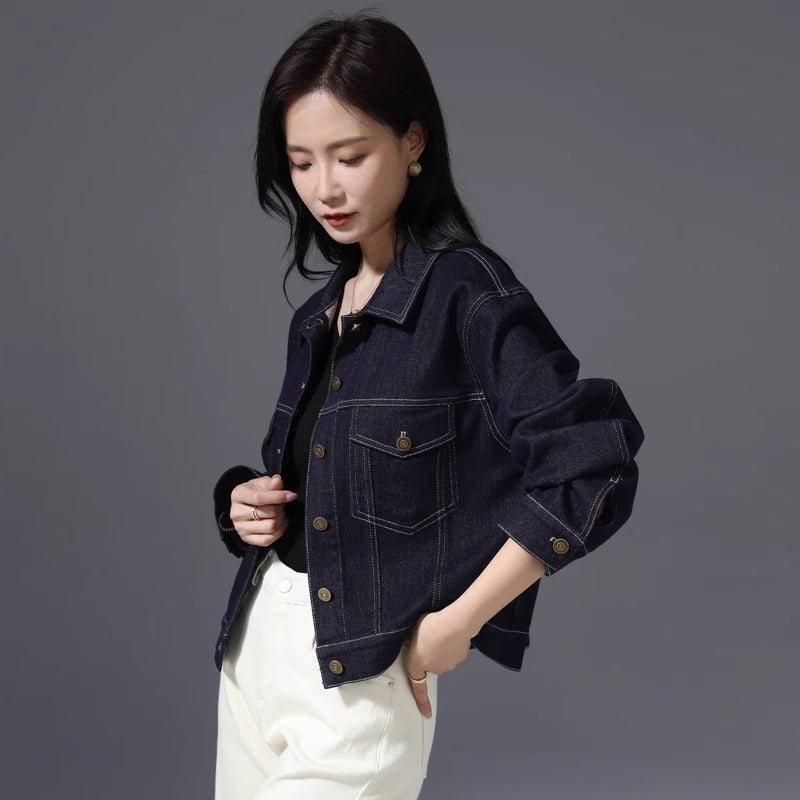 A woman wearing a dark indigo denim jacket with rolled-up sleeves and front flap pockets, paired with white pants, photographed against a solid dark gray studio background.
