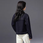 A woman shown from the back wearing a dark navy denim jacket and white pants, photographed against a solid dark gray studio background.